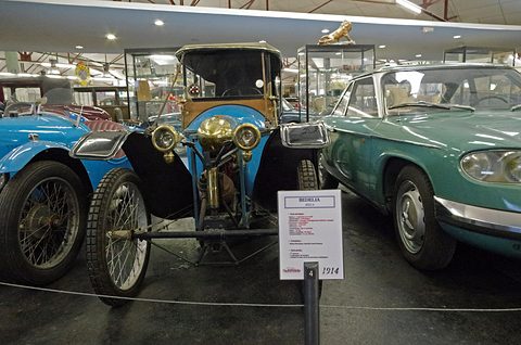 Blue narrow cyclecar with brass lamps between other cars, a museum label in front. Opens a larger view