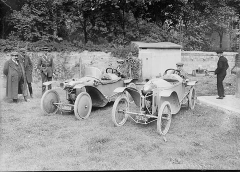 Two small cyclecars on grass before a garden wall, men in coats and caps standing around them. Opens a larger view