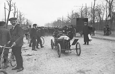 A small cyclecar on a cobbled avenue lined with bare trees, men with bicycles standing in the foreground.