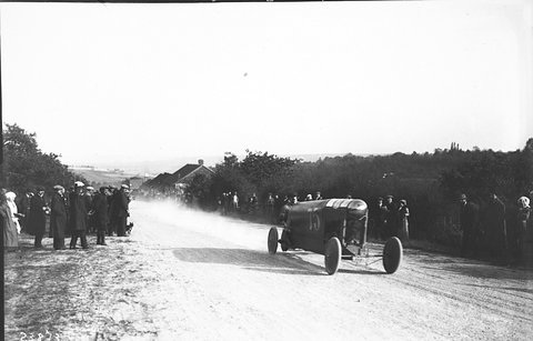 A small cyclecar raising dust on a pale road as spectators line the verge. Opens a larger view