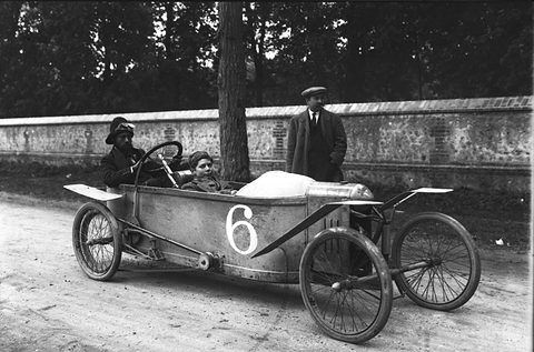 A long narrow cyclecar numbered 6 with wire wheels and cycle wings on a dirt road by a stone wall. Opens a larger view