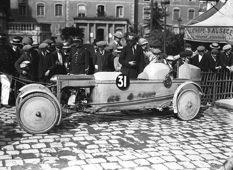 A low racing cyclecar numbered 31 with covered wheels on cobbles, a crowd of men in caps behind a rail.