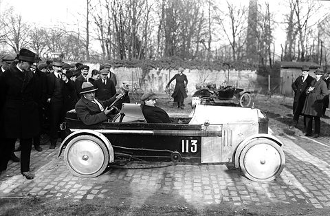 A low cyclecar numbered 113 with white disc wheels on cobbles, a crowd of men in hats behind. Opens a larger view