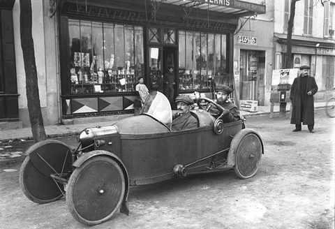 A narrow cyclecar with disc wheels outside a shop, one man seated behind another, a pedestrian on the pavement.