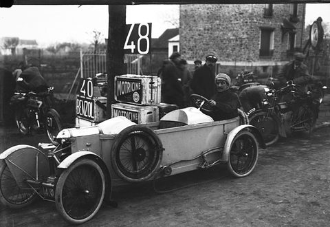 A pale cyclecar with fuel cans stacked behind it, parked among motorcycles and riders at a roadside control. Opens a larger view