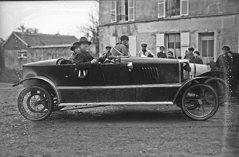 Side view of a long dark open cyclecar with wire wheels, spectators gathered by houses behind. Opens a larger view
