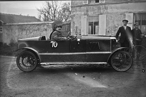 Side view of a long dark open cyclecar numbered 70 with wire wheels, men standing by a house behind. Opens a larger view