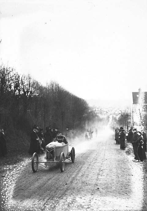 A small cyclecar driving up a dusty road between spectators, a hazy valley behind. Opens a larger view