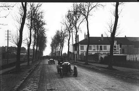 Two small cyclecars driving down a long cobbled road between bare trees and houses. Opens a larger view