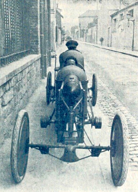 Rear view of a narrow cyclecar with two men seated in tandem, wire wheels and a belt-drive axle, on a walled street. Opens a larger view