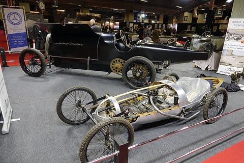 Bare cyclecar frame with a dented aluminium tub and wire wheels beside a long black racing car. Opens a larger view