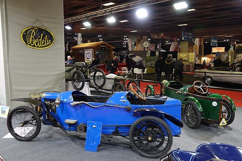 A bright blue racing cyclecar and a green cyclecar on display below an oval ‘Bédélia’ sign. Opens a larger view