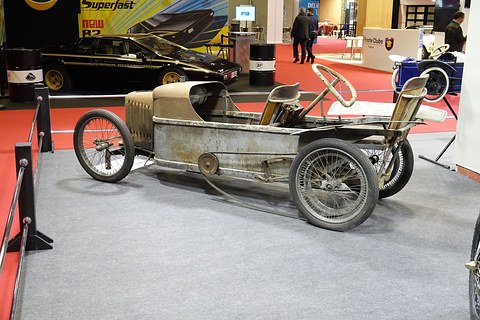 Side view of a bare weathered cyclecar chassis with two seats and wire wheels on grey carpet. Opens a larger view