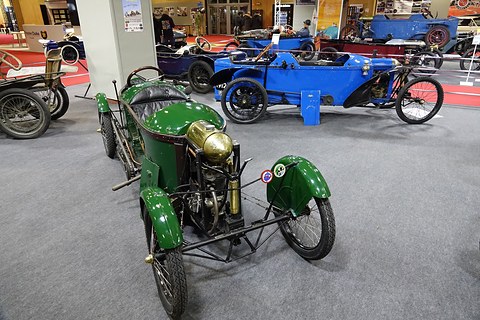 Green narrow cyclecar with cycle wings, brass engine parts and lamps, a blue racer behind. Opens a larger view