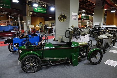 Green narrow cyclecar in front of several other small cyclecars in an exhibition hall. Opens a larger view