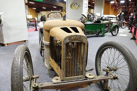 Front of a rusty cyclecar with a flat grille, a bare front axle and large wire wheels on carpet. Opens a larger view