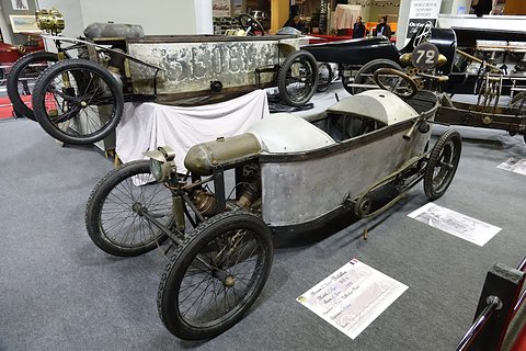 Weathered grey narrow cyclecar with wire wheels on carpet, other cyclecars on stands behind. Opens a larger view