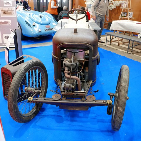 Front of a rusty narrow cyclecar with an exposed engine and wire wheels on a blue carpet. Opens a larger view