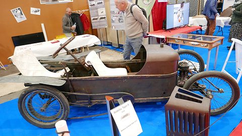Side view of a long low rusty cyclecar with wire wheels on a blue carpet, visitors behind. Opens a larger view