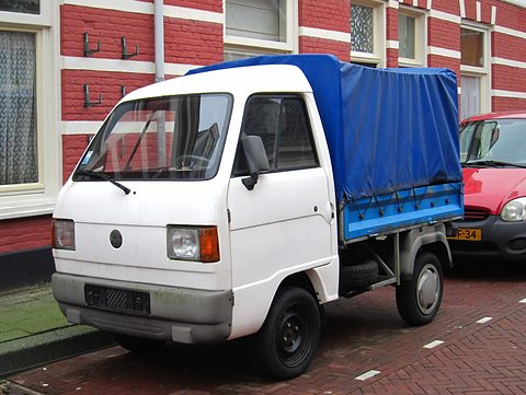 Small white cab-over pick-up truck with a blue canvas tilt over its load bed, parked before red-brick houses. Opens a larger view