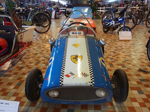 Front view of a small blue and white child-size racing car with a chequered stripe, a mesh grille and stickers. Opens a larger view