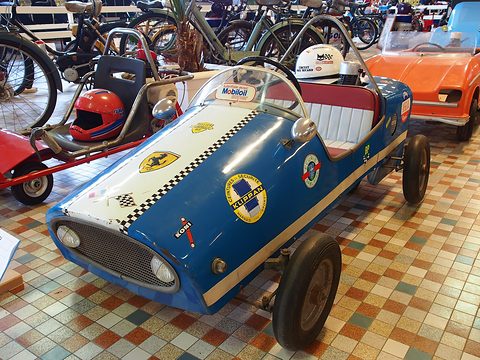 A small blue child’s racing car with a roll bar, a red helmet on its seat and Mobiloil and Ferrari stickers. Opens a larger view