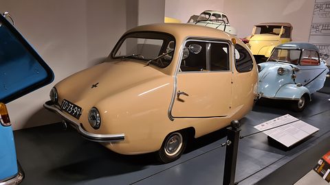 Rounded beige microcar on a dark plinth beside a pale blue bubble car, with other small cars behind.