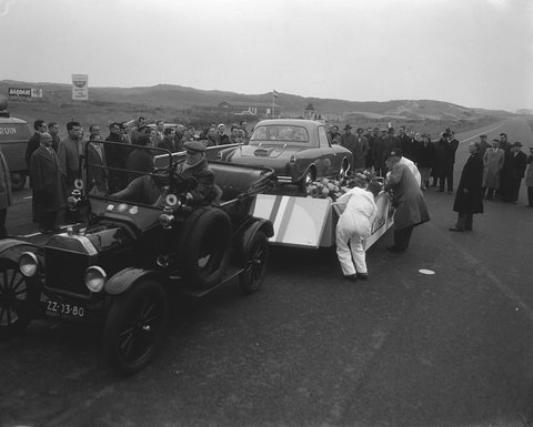 Black-and-white photograph of an old open car towing a trailer with a small coupé, a crowd and dunes behind.