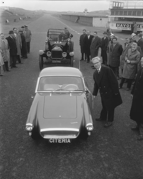 Black-and-white photograph of a small rounded sports coupé with CITERIA on its nose plate, a veteran car and onlookers behind.