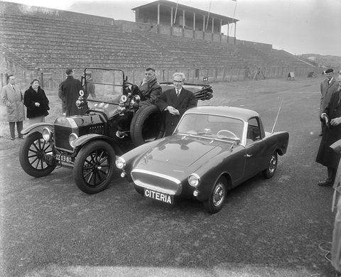 Black-and-white photograph of a low sports coupé parked beside a brass-era open tourer, men in overcoats around them, empty terraces behind. Opens a larger view