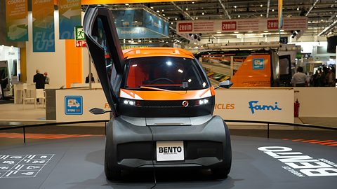 Front of a dark grey tiny car with orange trim and a BENTO plate, its door swung up, on a show stand. Opens a larger view