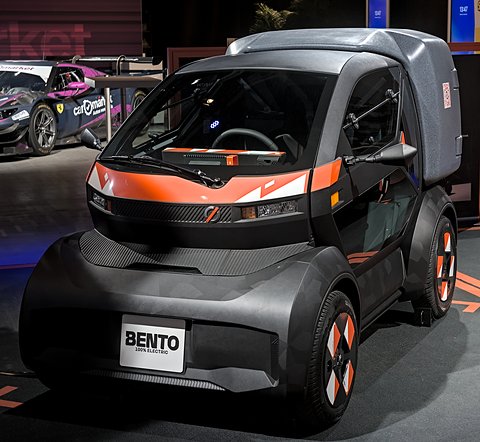 Dark grey tiny car with a large grey cargo box behind its single seat and a BENTO plate, on a show floor. Opens a larger view
