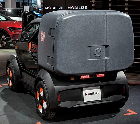 Rear of a tiny car with a large grey cargo box and a small tail-light strip, on a show floor. Opens a larger view