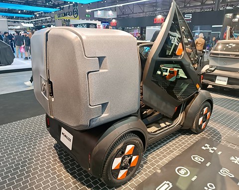Tiny car with a large grey cargo box and its door raised, on a patterned show floor, visitors behind. Opens a larger view