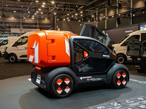 Side view of a tiny car with an orange cargo box and chequered wheel covers, white vans behind. Opens a larger view