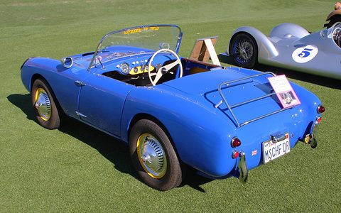 Blue open two-seater from behind on a lawn, a chrome luggage rack on the tail. Opens a larger view