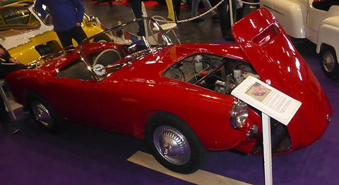 Red open sports car with its bonnet raised on a dark show carpet, a caption stand beside. Opens a larger view