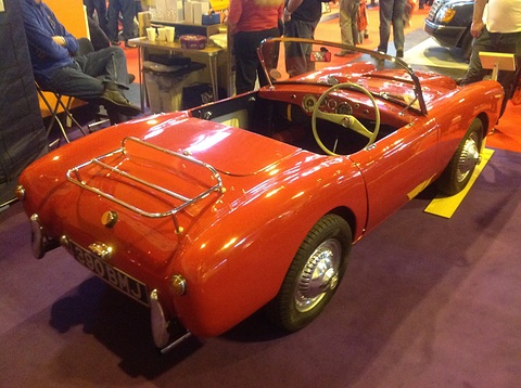 Red open sports car with a chrome boot rack seen from behind on a show carpet. Opens a larger view