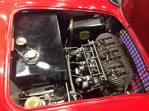 A finned three-cylinder engine with three carburettors next to a black fuel tank inside a red glass-fibre body. Opens a larger view