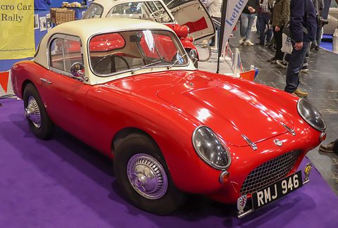 A tiny red sports car with faired-in headlamps, a mesh grille and a cream hardtop on a purple show carpet. Opens a larger view