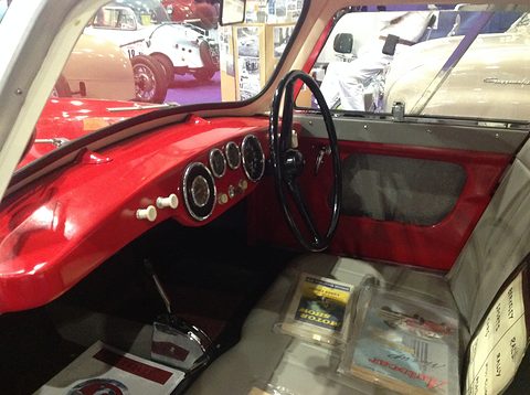 A red dashboard with four round dials and a thin black steering wheel, a gear lever rising from the floor. Opens a larger view