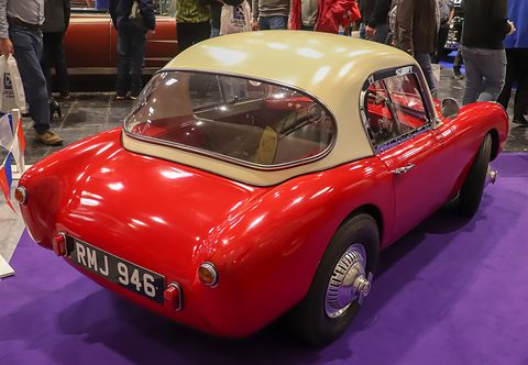 Rear three-quarter view of a small red sports car with a cream hardtop, round tail lamps and chrome hubcaps. Opens a larger view