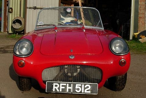 Red open two-seater seen from the front with a mesh grille and round headlamps, a shed behind. Opens a larger view