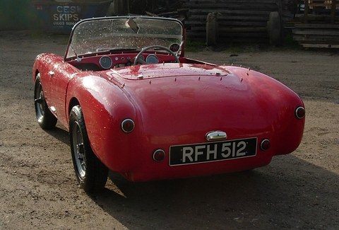 Red open two-seater seen from behind on a gravel yard. Opens a larger view