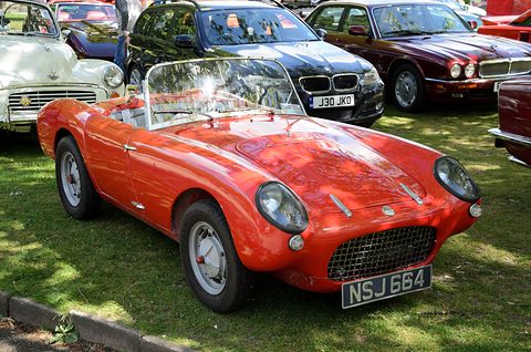 Orange-red open two-seater on grass among modern cars, seen from the front left. Opens a larger view