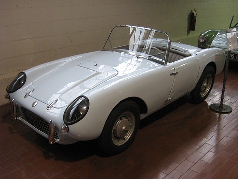 White open two-seater on a brick floor in a museum, seen from the front left. Opens a larger view