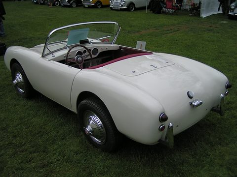 White open two-seater with red seats seen from behind on a lawn. Opens a larger view