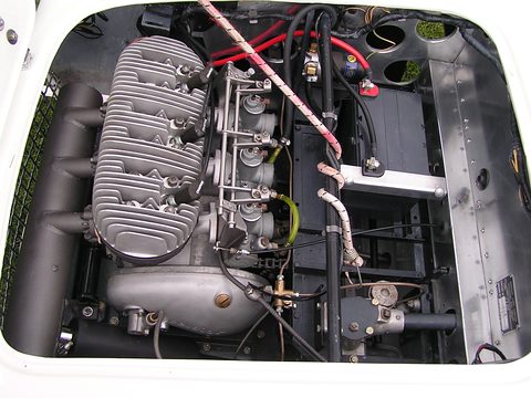 Finned three-cylinder engine with three carburettors inside a white engine bay. Opens a larger view