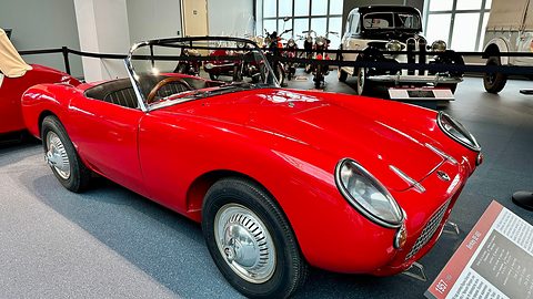 Red open two-seater on a grey museum floor behind a rope, seen from the front right. Opens a larger view