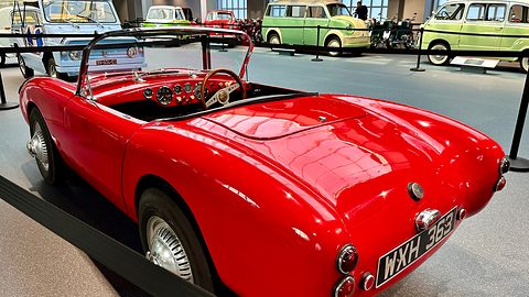 Red open two-seater from the rear left in a museum, other small cars in the background. Opens a larger view